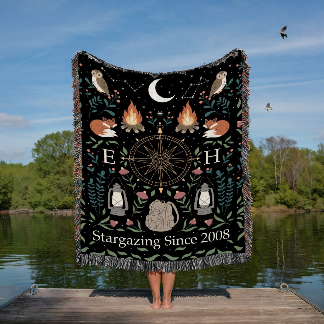 Person holding a decorative blanket with nature-themed designs by a lake.