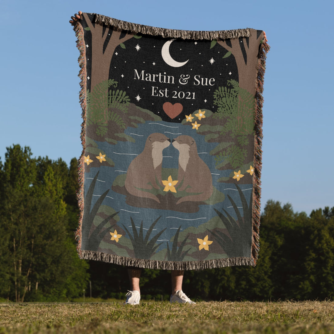 Person holding a blanket with a design of two otters and text 'Martin & Sue Est 2021' against a natural background.