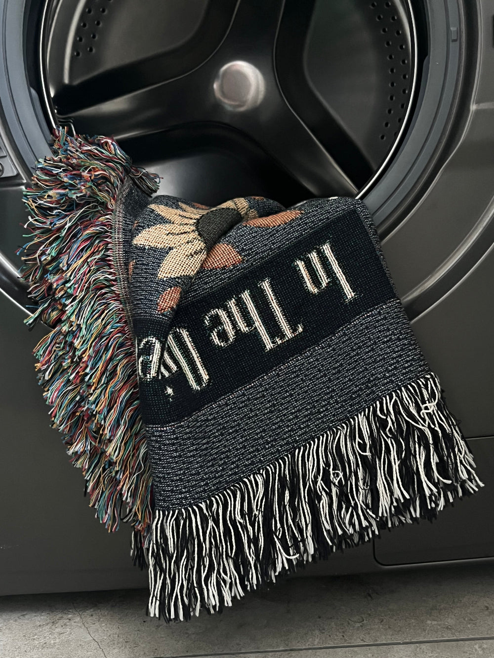 Decorative cotton throw blanket with fringe draped over an open washing machine